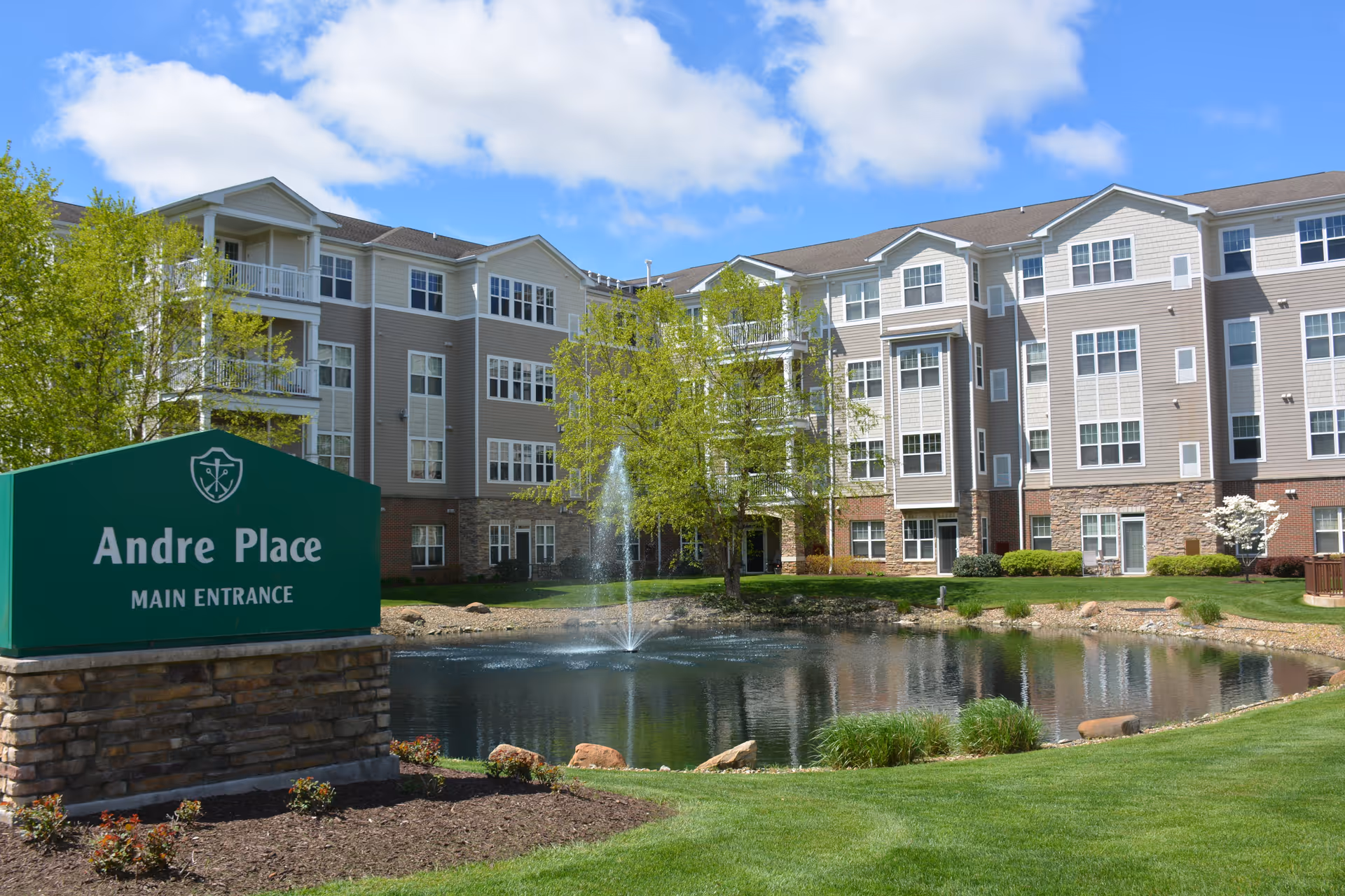 Exterior view of a multi-story senior living building named Andre Place with a pond and a fountain in front, surrounded by green grass and trees under a partly cloudy blue sky.