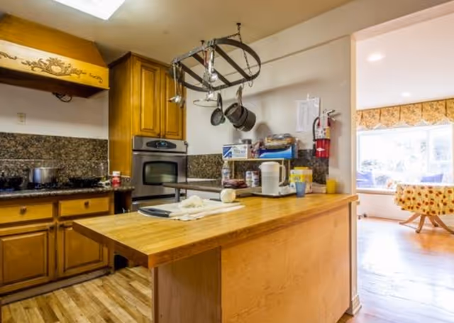 Wood-furnished kitchen with a central island, hanging pot rack, oven and stovetop, and countertop appliances opening into a sunny dining area.