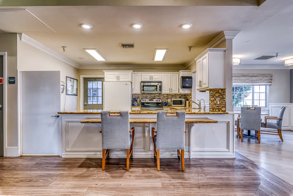 A bright and clean kitchen area in a senior living facility with white cabinets, a refrigerator, microwave, stove, and a granite countertop island with two gray cushioned chairs. To the right, there is a dining area with a table and chairs near a window with blinds.