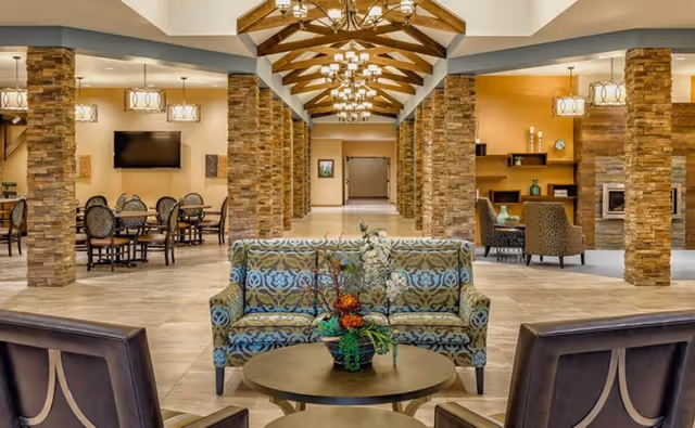 Spacious lobby-style common area with patterned sofas and chairs arranged around round tables, stone columns, and decorative chandeliers.