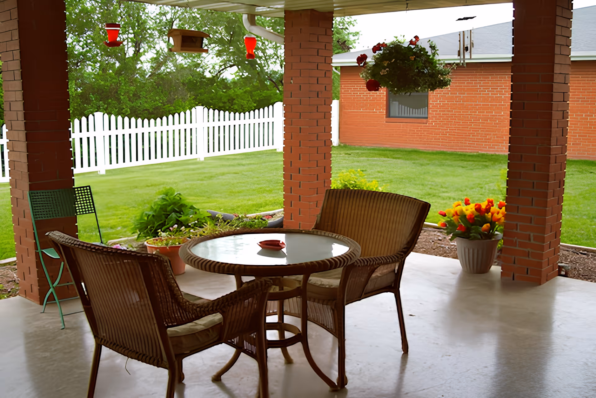 Covered patio with a round table and wicker chairs, brick columns, hanging plants and a white picket fence overlooking a lawn.