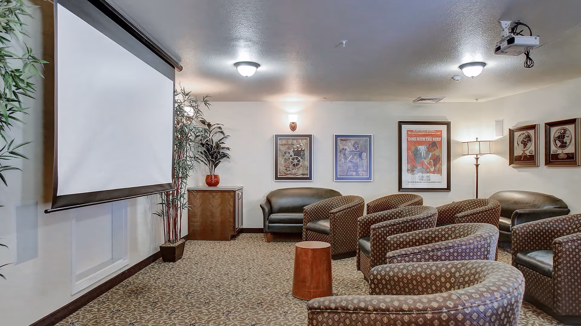 A cozy media room with patterned armchairs and leather sofas arranged facing a large pull-down projector screen. The room has framed artwork on the walls, a floor lamp, and potted plants, creating a comfortable and inviting atmosphere.