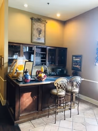 A small indoor bar area with a dark wooden counter and two high cushioned bar stools with decorative backs. Behind the counter is a glass-front cabinet with shelves holding glasses and other items. The walls are painted beige with framed artwork hanging, and the floor is tiled with a light pattern. The space is softly lit by ceiling lights.