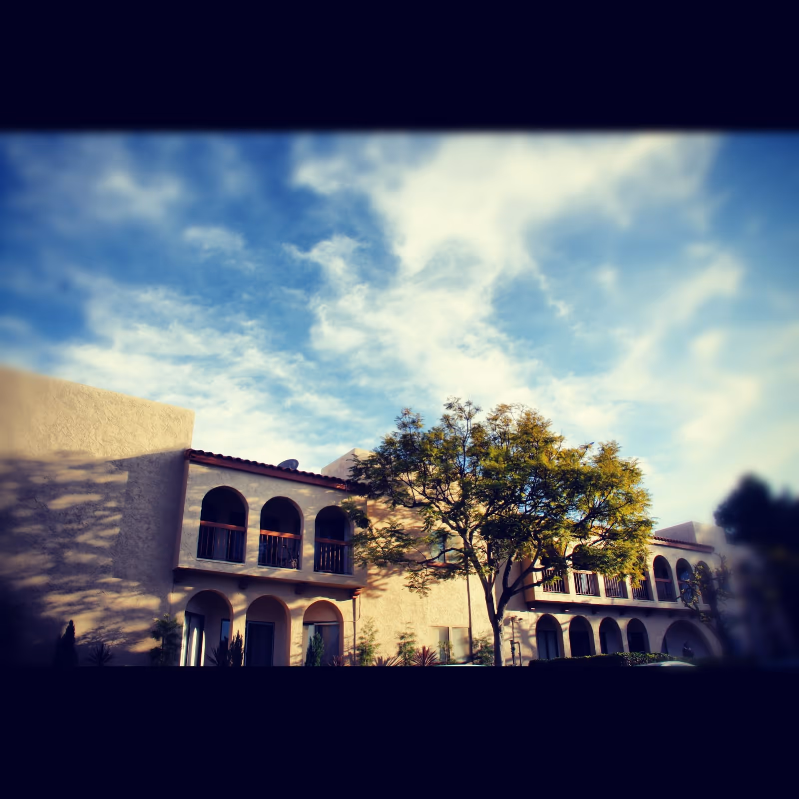 Exterior view of a two-story building with arched balconies and windows, a tree in front, and a partly cloudy blue sky above.