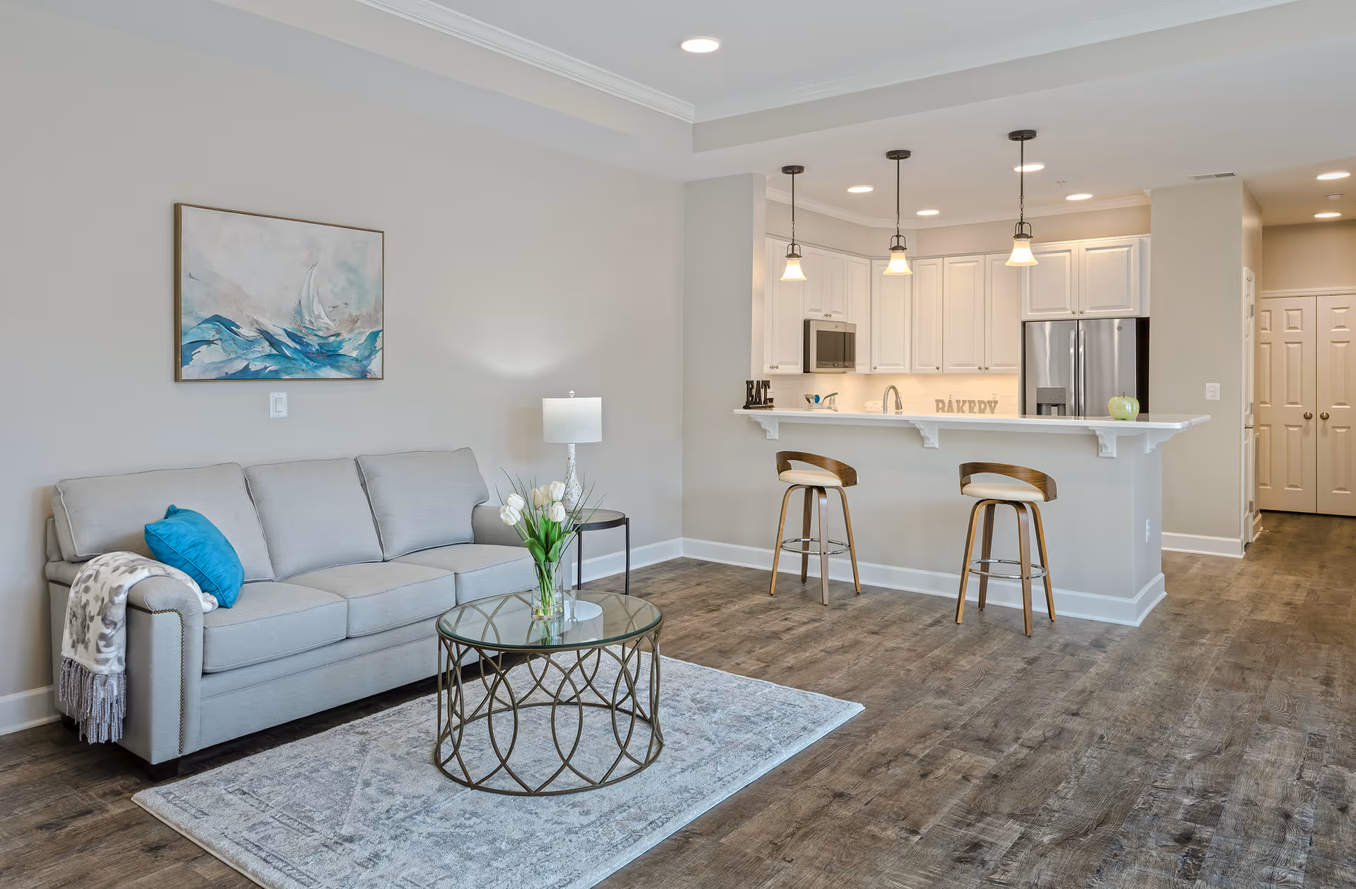 Open-concept living room with a gray sofa, glass coffee table, and a kitchen bar with two stools and pendant lights.