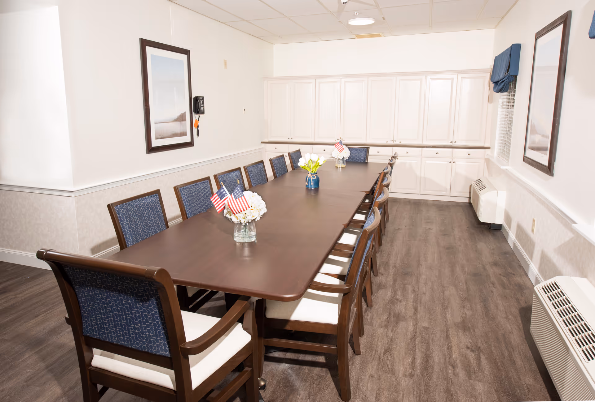 A long rectangular dining table with twelve wooden chairs featuring blue cushioned backs and white seats. The table has small flower vases with American flags as decoration. The room has white walls, two framed pictures, a wall-mounted phone, white cabinetry along the back wall, and wood-style flooring.