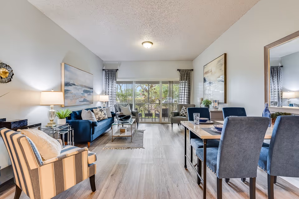 A bright and spacious senior living facility interior featuring a living room and dining area. The living room has a blue sofa with patterned cushions, a striped armchair, a glass coffee table, and two side tables with lamps. The dining area includes a table set with plates and glasses, surrounded by four blue upholstered chairs. Large sliding glass doors at the back open to an outdoor patio with greenery visible outside. The walls are decorated with framed artwork and a large mirror.
