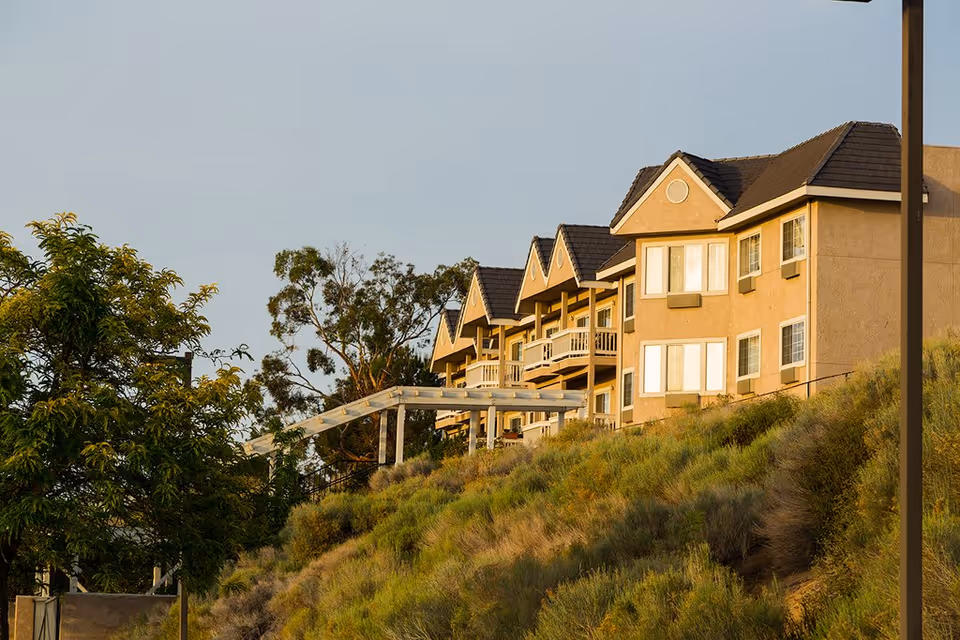 Row of beige multi-story assisted living buildings on a grassy hillside under a clear sky.