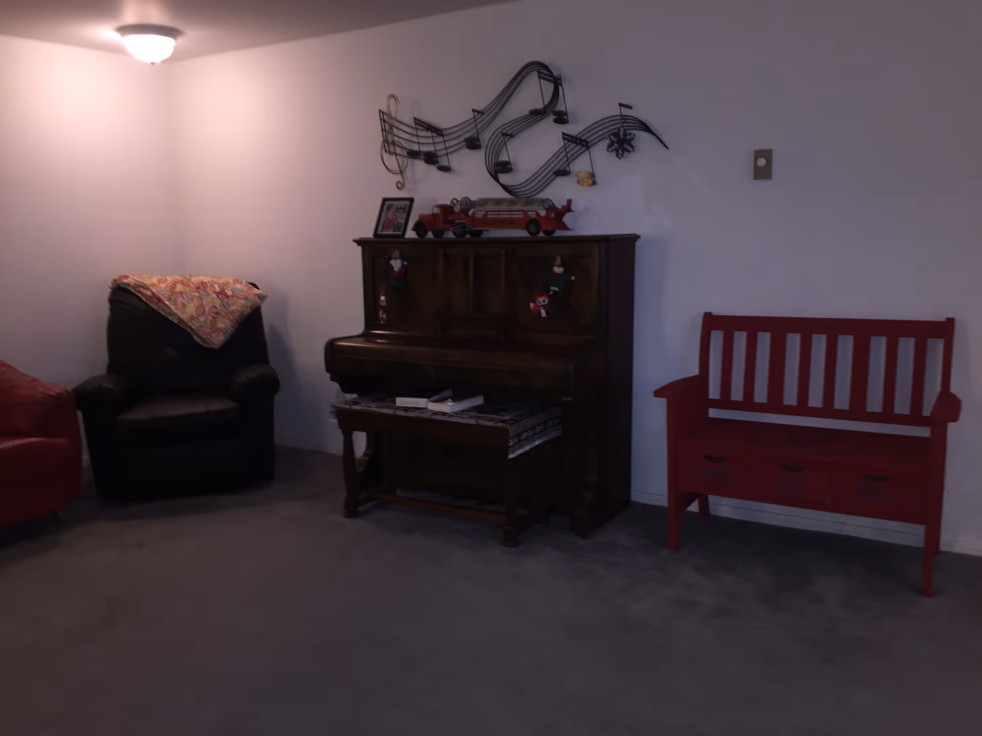 Living room with an upright piano against a wall, a black armchair to the left and a red bench to the right under a musical notes wall decoration.