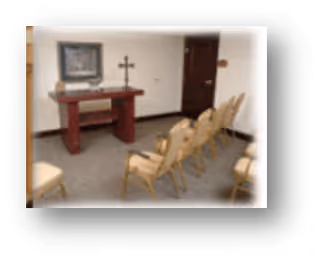 A small room with several beige chairs arranged in rows facing a wooden table with a framed picture and a cross on it, and a closed wooden door in the background.