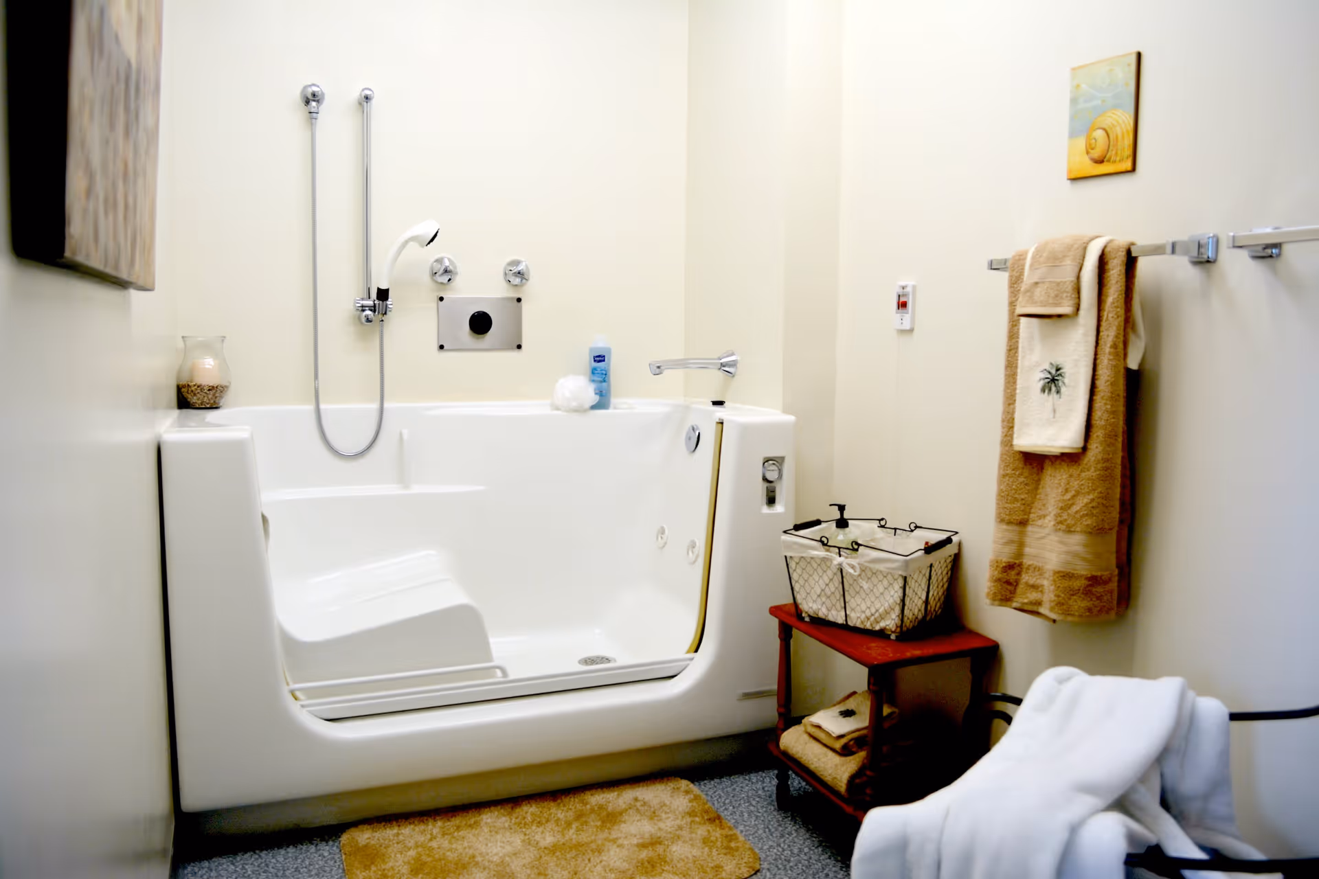 A clean, accessible bathroom featuring a walk-in bathtub with a built-in seat and handheld showerhead. The room has beige walls, a small wooden shelf holding a basket and folded towels, a towel rack with beige towels, a bath mat on the floor, and a white robe hanging on a chair.
