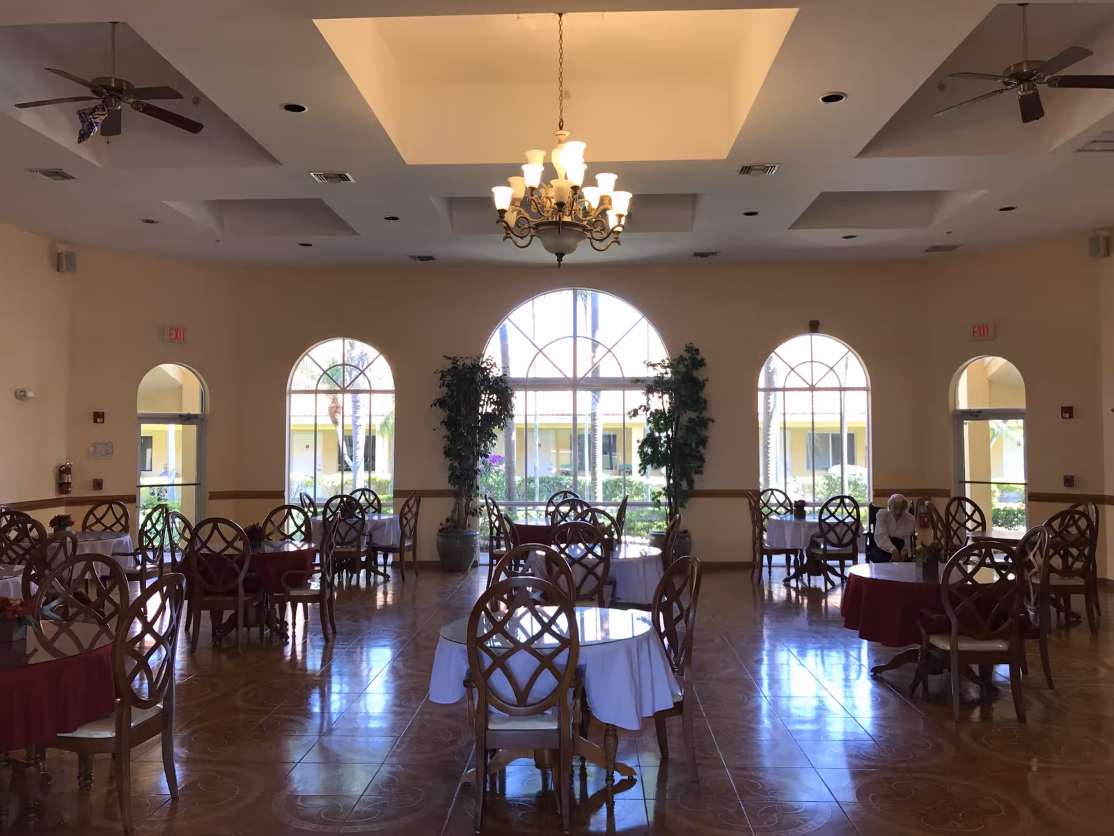 A spacious dining room with several round tables covered with white and red tablecloths, surrounded by wooden chairs. Large arched windows let in natural light and show a view of palm trees and a courtyard outside. Two ceiling fans and a chandelier hang from the ceiling. A single elderly person is seated at a table on the right side of the room.