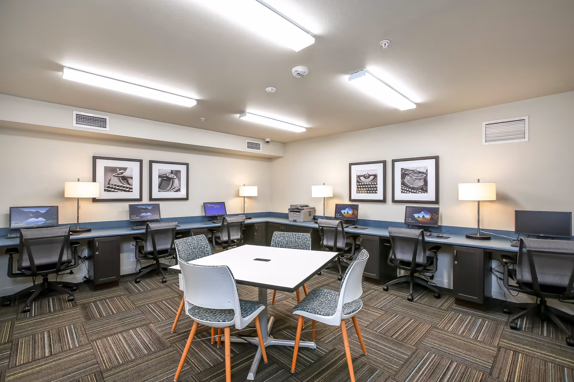 A computer room with multiple workstations along two walls, each equipped with a computer monitor and office chair. There are three table lamps evenly spaced on the desks. In the center of the room is a square table with four chairs around it. The walls are decorated with framed black and white photographs, and the room is well-lit with ceiling lights.