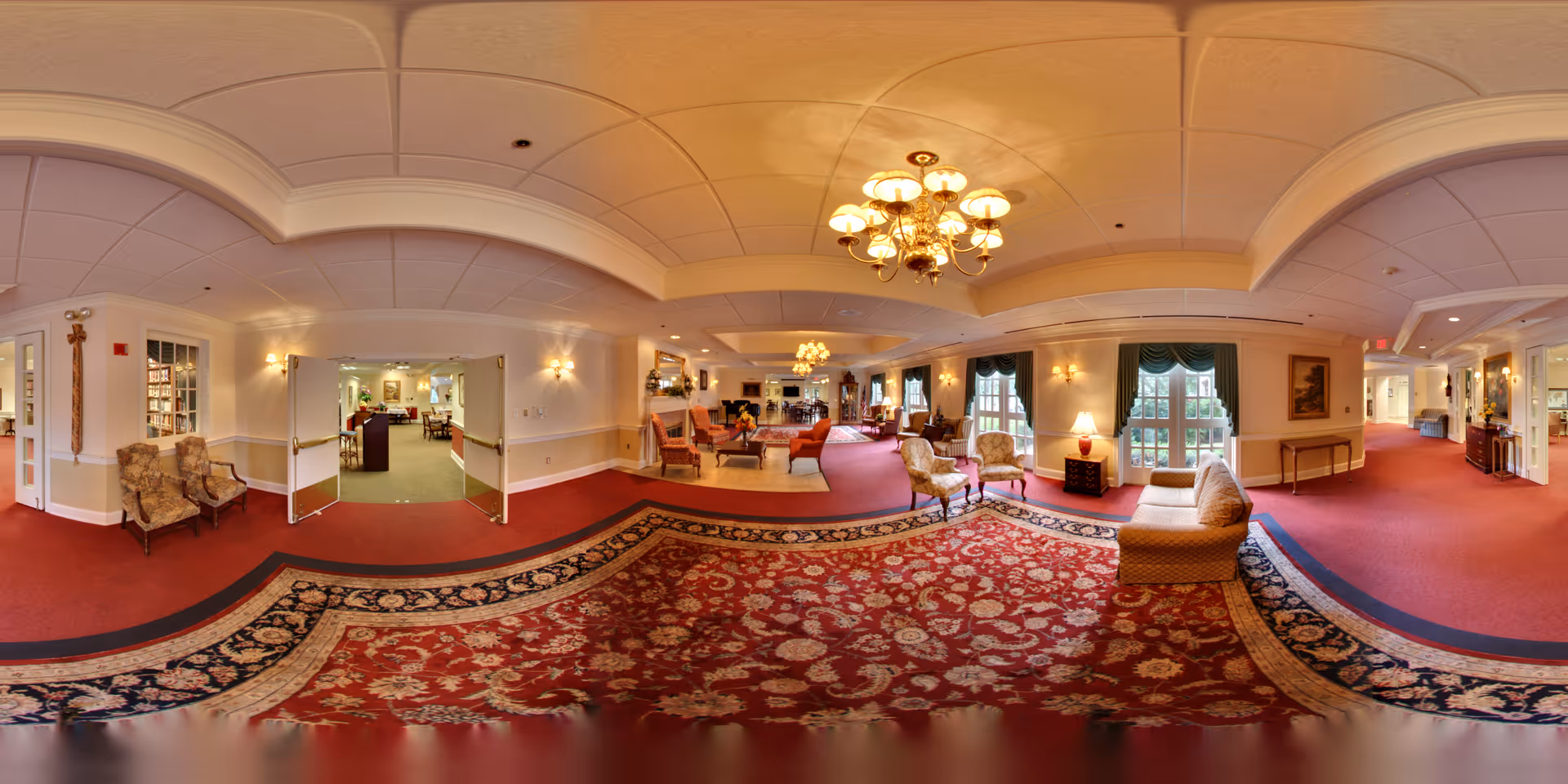 A spacious and elegantly decorated senior living facility common area with red carpet and a large patterned rug. The room features multiple seating arrangements including armchairs and sofas, chandeliers hanging from a white paneled ceiling, wall sconces, large windows with green drapes, and a hallway leading to other rooms.