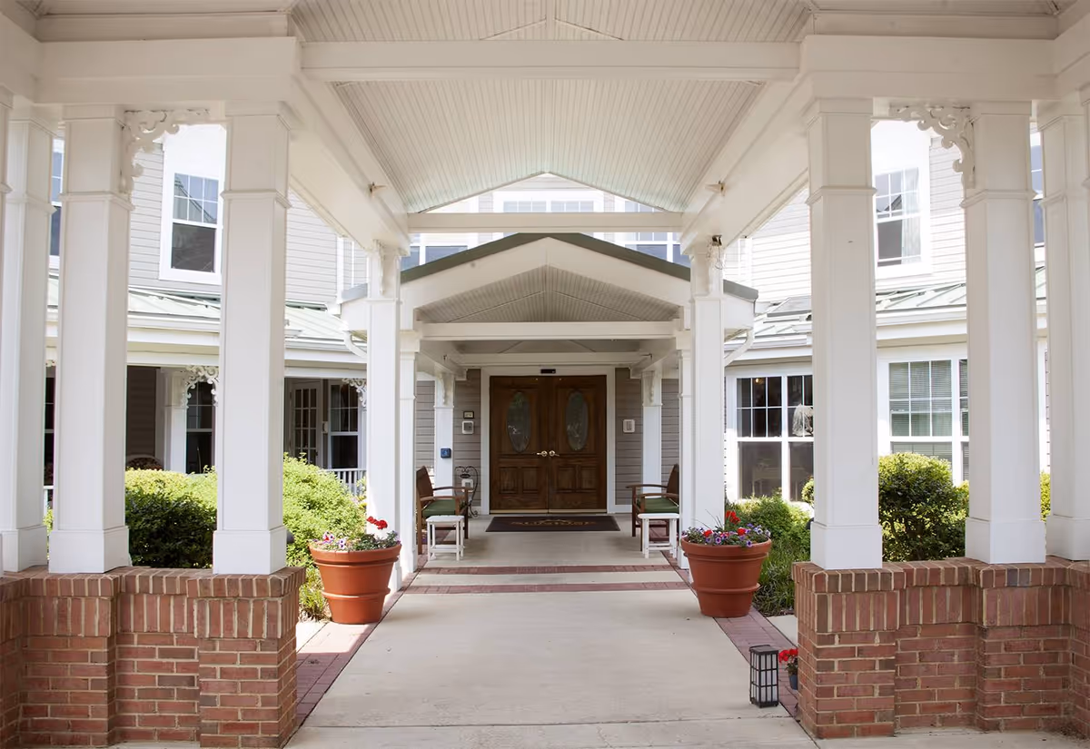 Covered entrance to a senior living facility with white columns, brick bases, potted flowers, benches, and double wooden doors under a peaked roof.