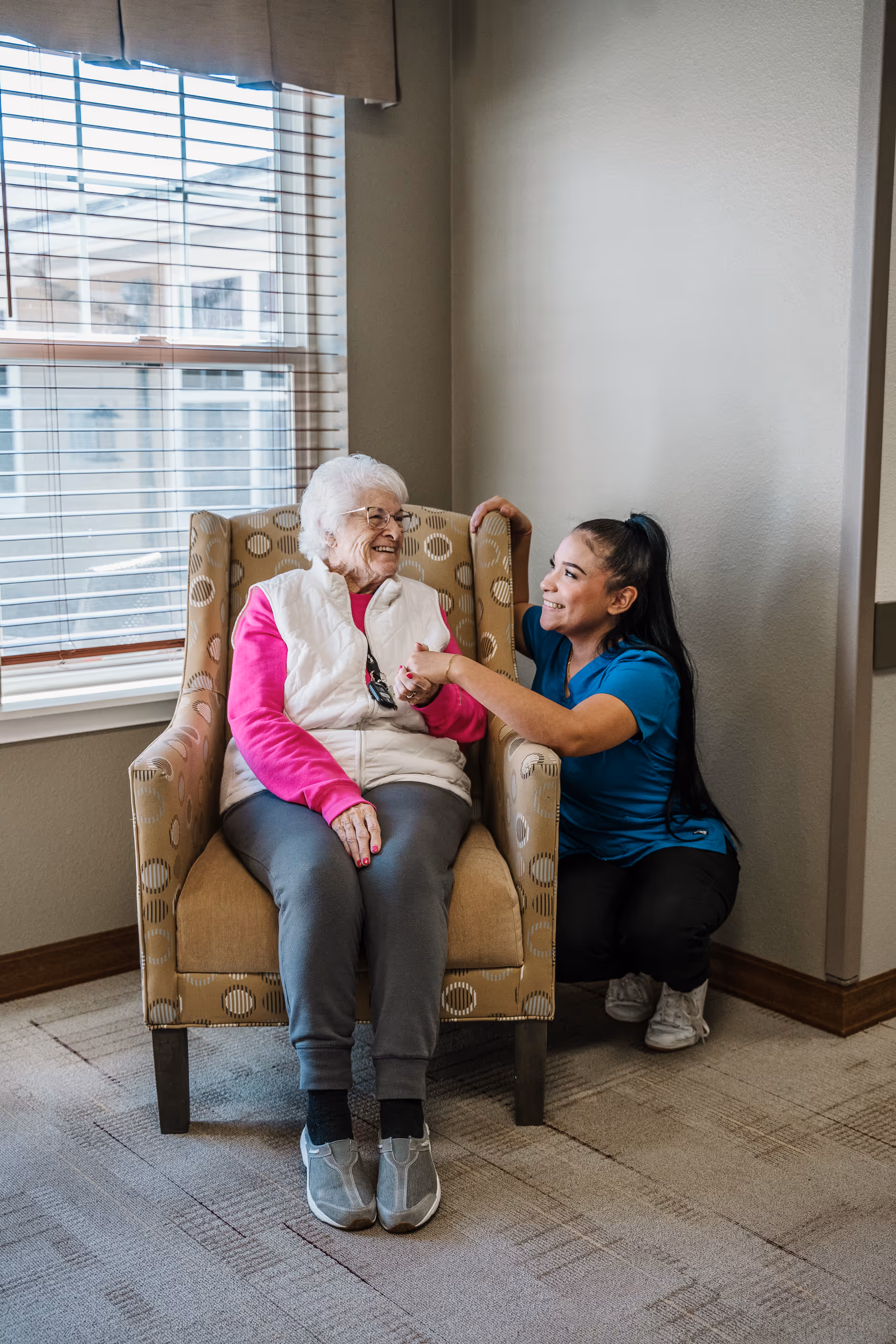 An elderly woman with white hair wearing a white vest and pink long-sleeve shirt sits in a patterned armchair near a window with blinds. A younger woman in a blue shirt and black pants kneels beside her, smiling and holding the elderly woman's hand in a supportive and caring manner. The setting appears to be a cozy indoor space with beige walls and carpeted floor.