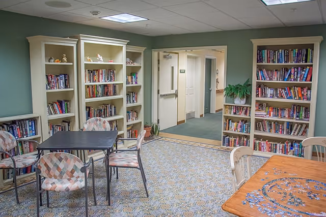 A cozy common area with bookshelves filled with books along the walls, several chairs with patterned upholstery around a black table, and a wooden table with a partially completed jigsaw puzzle. The room has a carpeted floor and a doorway leading to a hallway.
