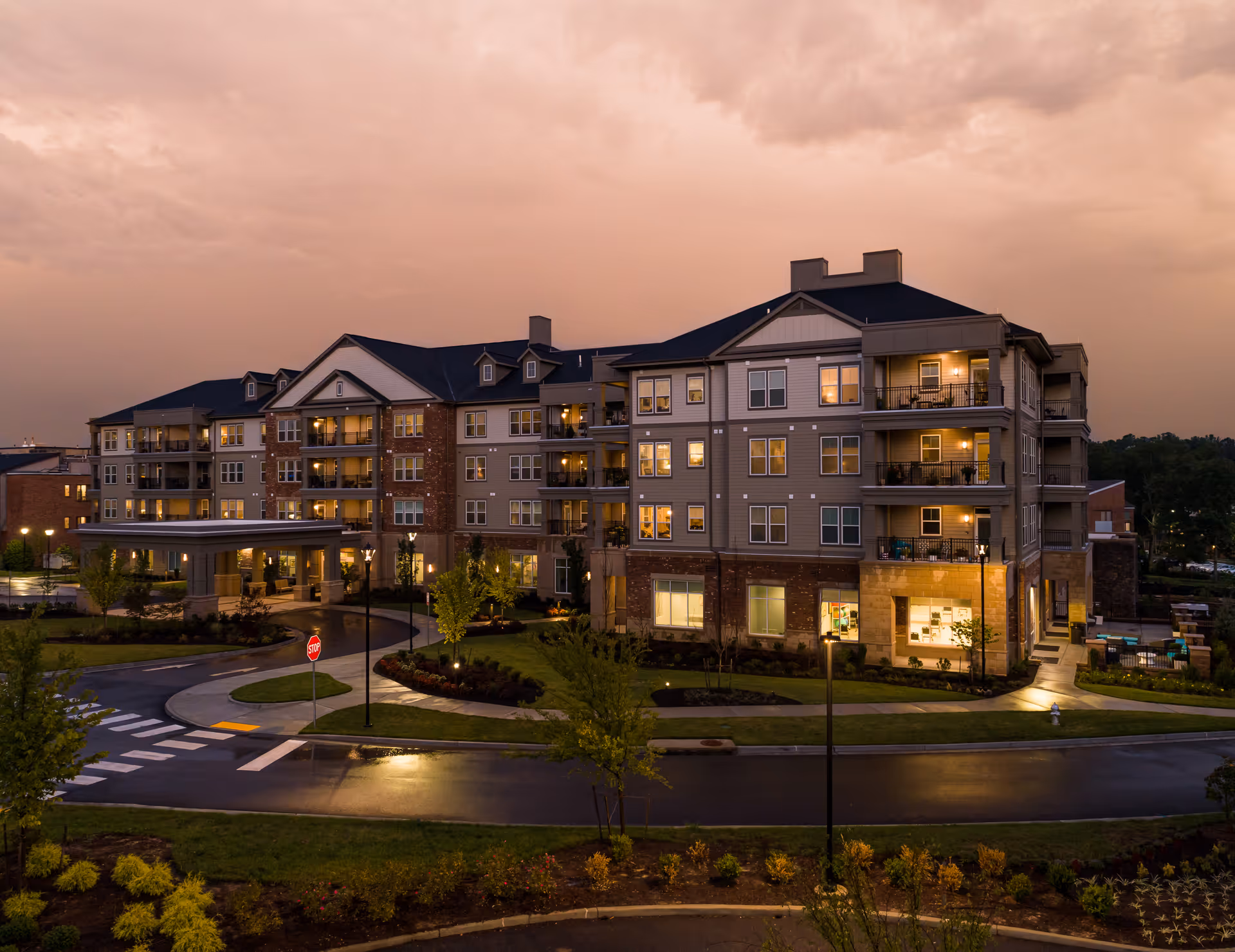 Exterior view of a multi-story senior living facility building at dusk with lights on inside and landscaped surroundings including trees and bushes.