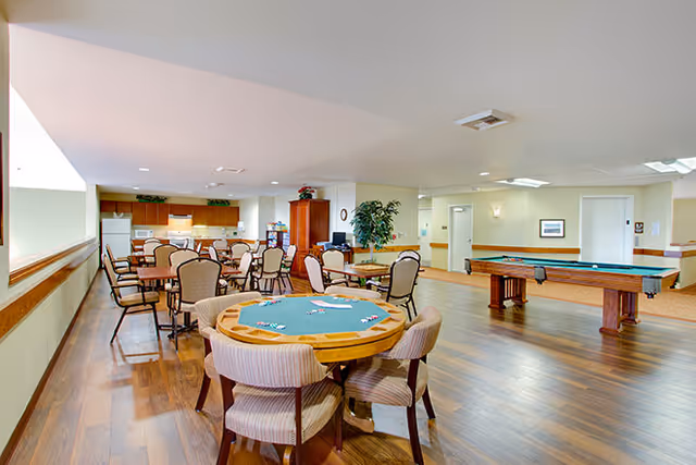 A spacious common area in a senior living facility featuring a round card table with four chairs in the foreground, multiple dining tables with chairs, a pool table, and a kitchenette with cabinets, refrigerator, and microwave in the background. The room has wooden flooring, light-colored walls, and ceiling lights.