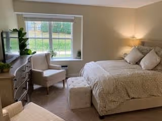 A cozy bedroom with a large bed covered in beige bedding and multiple pillows. There is a cushioned ottoman at the foot of the bed, a beige armchair next to a window with white blinds, and a wooden dresser with a TV on top. A potted plant is placed on the dresser, and a lamp on a nightstand provides warm lighting.