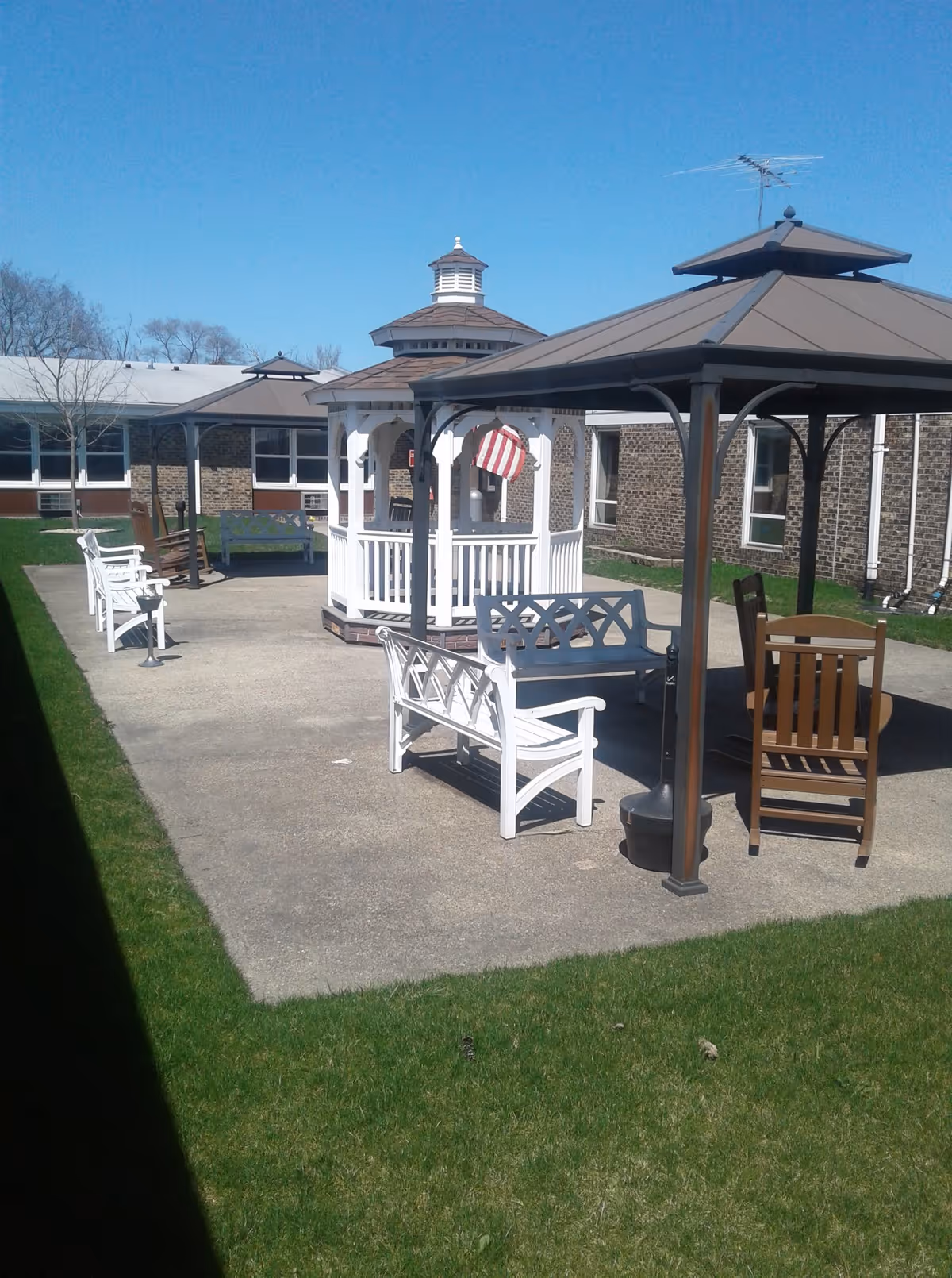 Outdoor patio area with white benches, a wooden chair, two gazebos with brown roofs, and a white gazebo with an American flag hanging inside. The area is surrounded by a brick building and green grass under a clear blue sky.