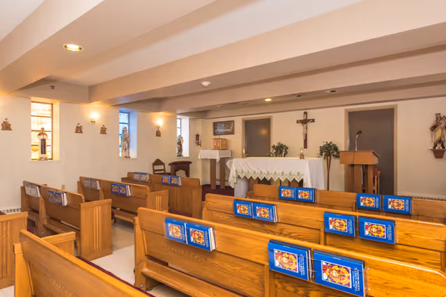 Interior view of a small chapel with wooden pews arranged in rows, each holding blue hymnals. At the front is an altar covered with a white cloth, a crucifix on the wall behind it, and religious statues and decorations along the walls.