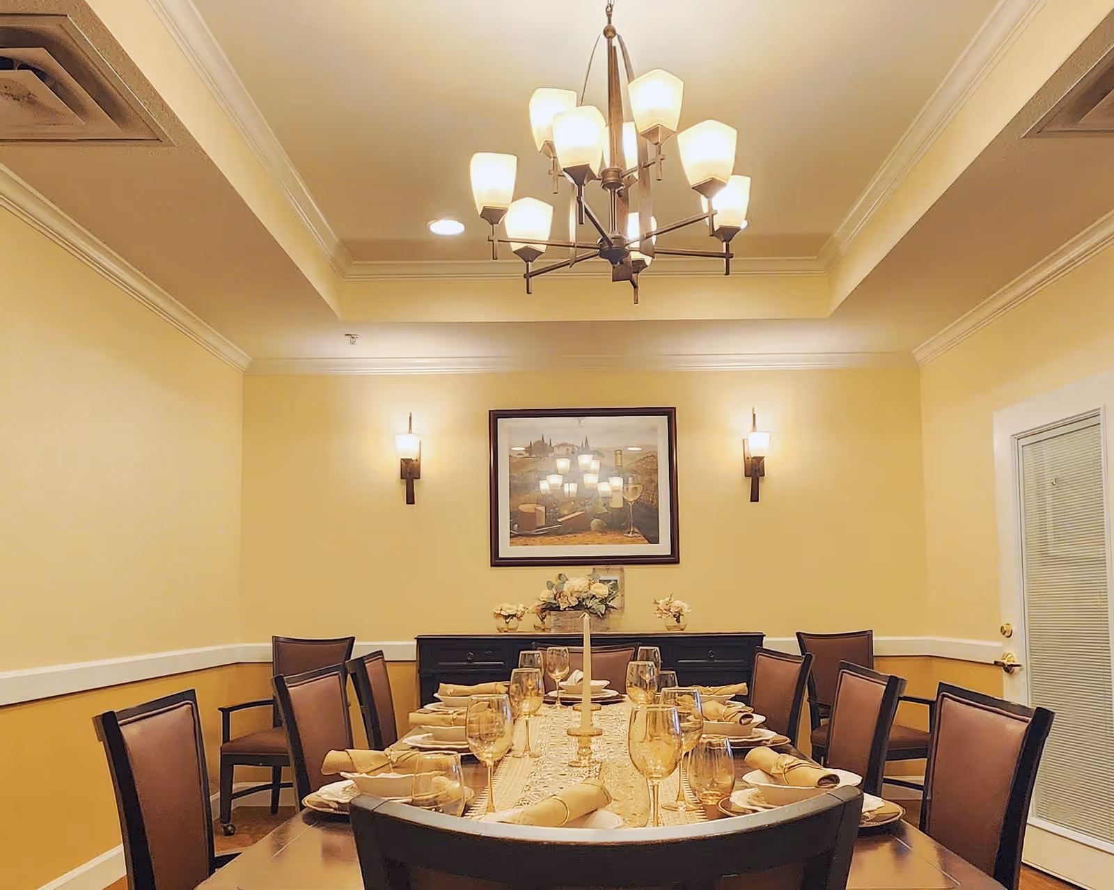 A formal dining room with a long wooden table set for a meal, featuring plates, rolled napkins, and wine glasses. The room has beige walls, a chandelier with multiple lights hanging from the ceiling, two wall sconces, a framed painting on the wall, and a sideboard with floral decorations. There is a door with a window on the right side.