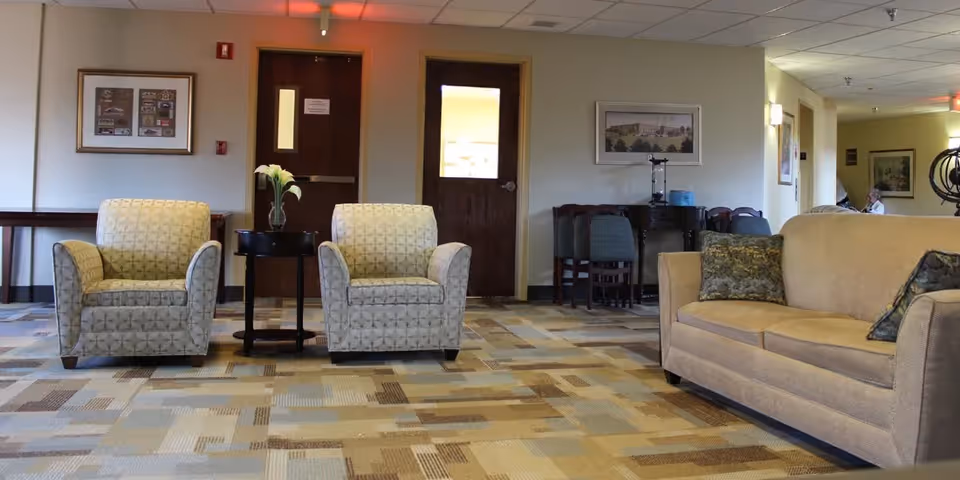 A seating area in a nursing and rehabilitation center with two patterned armchairs and a beige sofa with decorative pillows. A small round table with a vase of flowers is placed between the armchairs. The room has a patterned carpet, framed pictures on the walls, and wooden doors in the background.