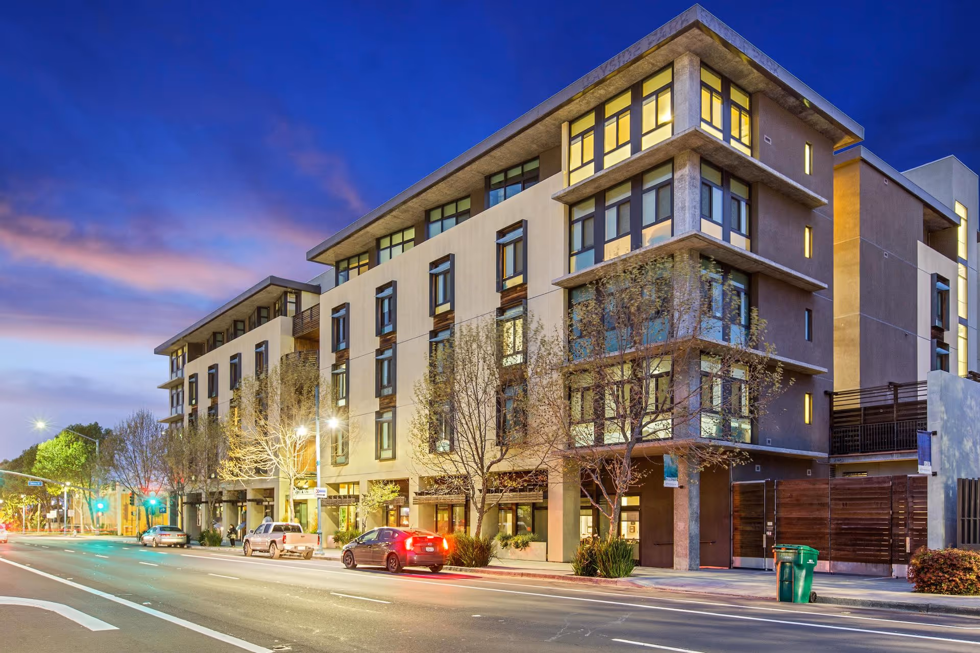 A modern four-story senior living building with illuminated windows and street traffic at dusk.