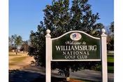 A green and white sign that reads 'Welcome to Williamsburg National Golf Club' with trees and a clear blue sky in the background.