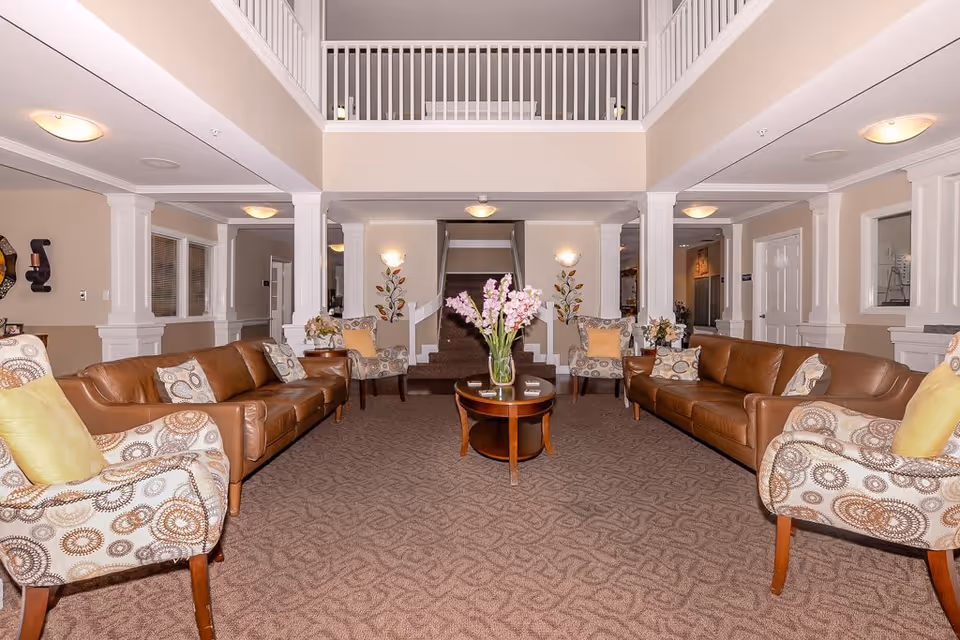 A spacious and well-lit living room area with two brown leather sofas and four patterned armchairs arranged around a wooden coffee table with a vase of pink flowers. The room features beige walls, carpeted flooring, white pillars, and a staircase in the background leading to an upper level with a white railing.