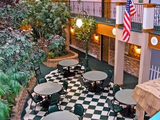 Indoor courtyard area with round tables and chairs arranged on a black and white checkered floor. There are large green plants and trees, brick walls, and an American flag hanging from the upper level. The space is well-lit with wall-mounted lights and a streetlamp-style light fixture.