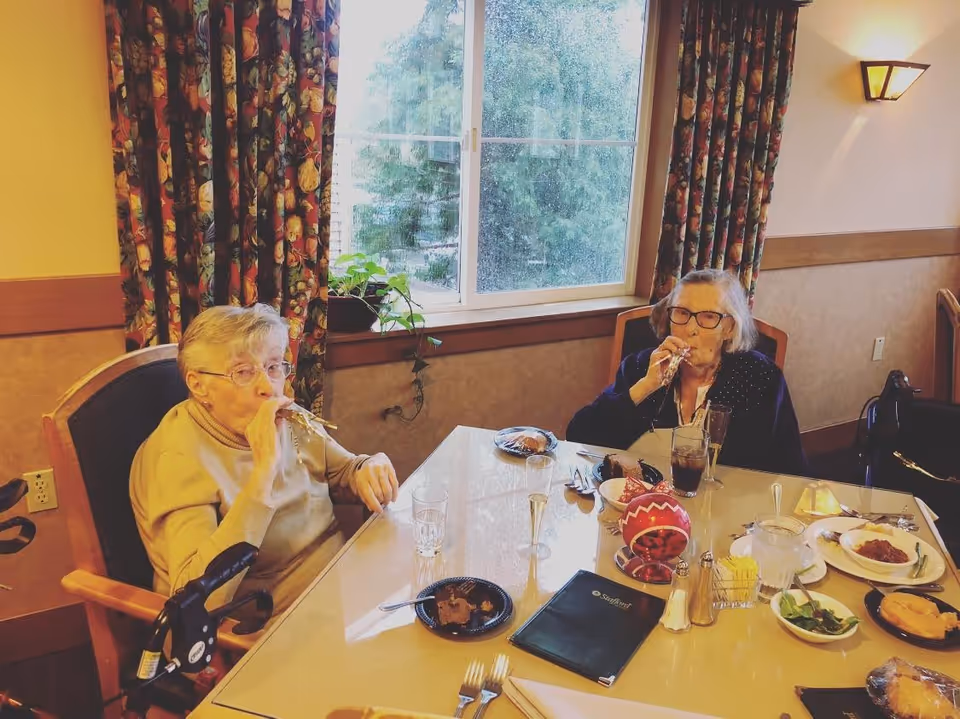 Two elderly women sitting at a dining table in a room with floral curtains and a large window. They are blowing party blowers and have plates of food, drinks, and utensils on the table in front of them.