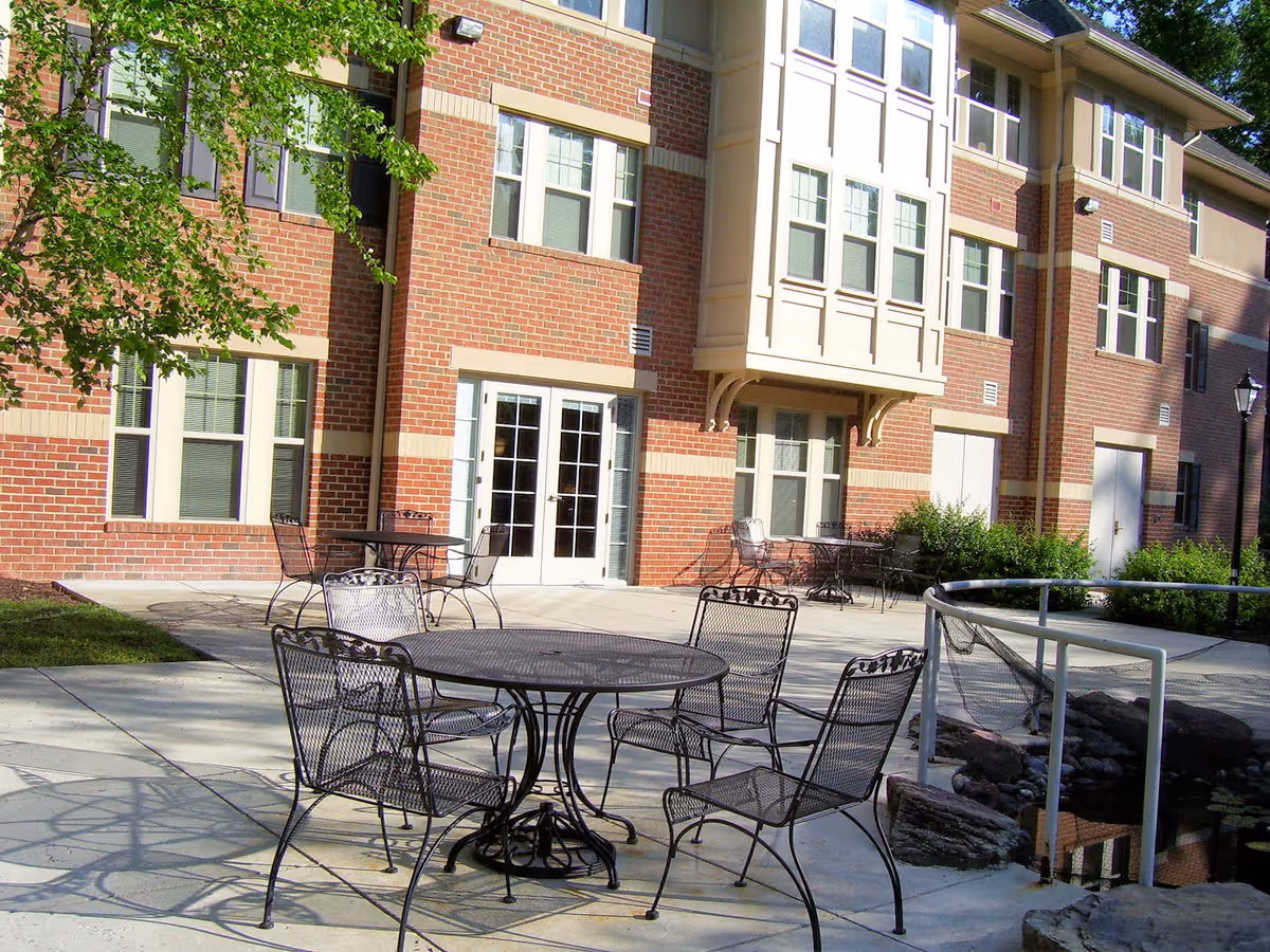 Outdoor patio area at Victory Terrace (Seniors 62+) with metal tables and chairs on a concrete surface, adjacent to a brick building with multiple windows and a glass door. There is a small pond with rocks and a white railing nearby, and some greenery including bushes and a tree.