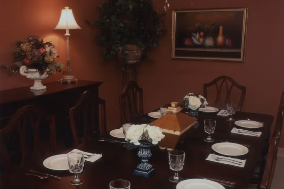 A formal dining room with a dark wooden table set for six people with white plates, crystal glasses, and silverware. The table is decorated with two small floral arrangements and a wooden decorative box in the center. In the background, there is a sideboard with a lamp and a floral arrangement, a potted plant, and a framed painting on the wall.