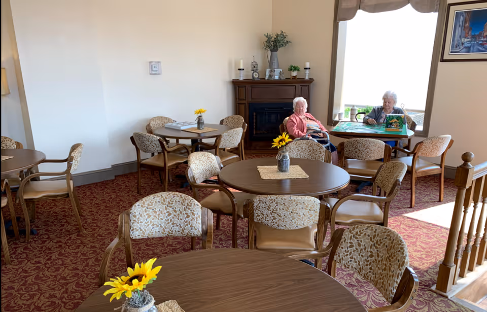 A cozy common area in a senior living facility with round wooden tables and patterned chairs. Two elderly women are seated near a large window, one sitting in a wheelchair and the other at a table playing a board game. The room has a red patterned carpet, a wooden fireplace mantel with decorative items, and small vases with sunflowers on the tables.