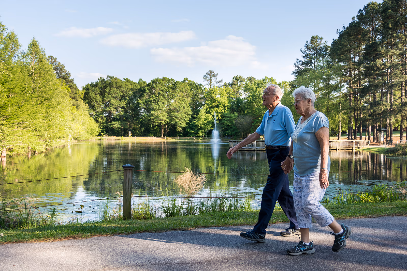 An elderly couple walking hand in hand along a paved path next to a pond with a fountain, surrounded by lush green trees under a partly cloudy sky.