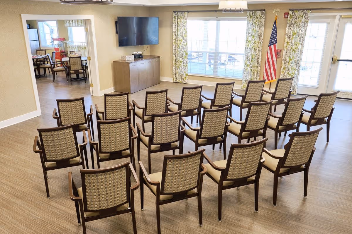 A meeting or presentation room with rows of wooden chairs facing a wall-mounted flat screen TV. The room has large windows with floral curtains and an American flag in the corner. Adjacent to the room is a dining area with tables and chairs visible through an open doorway.