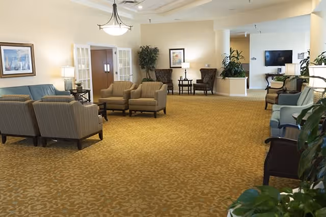 A spacious senior living facility common area with beige patterned carpet, multiple seating arrangements including armchairs and sofas, side tables with lamps, framed artwork on the walls, and several potted plants. The room is well-lit with ceiling lights and natural light from windows.