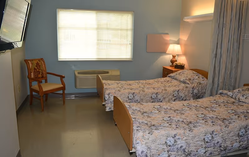 A room with two single beds covered in floral bedspreads, a wooden chair with a cushioned seat and backrest, a window with blinds, a wall-mounted air conditioning unit, a nightstand with a lamp and some decorative items, and a curtain partially visible on the right side.