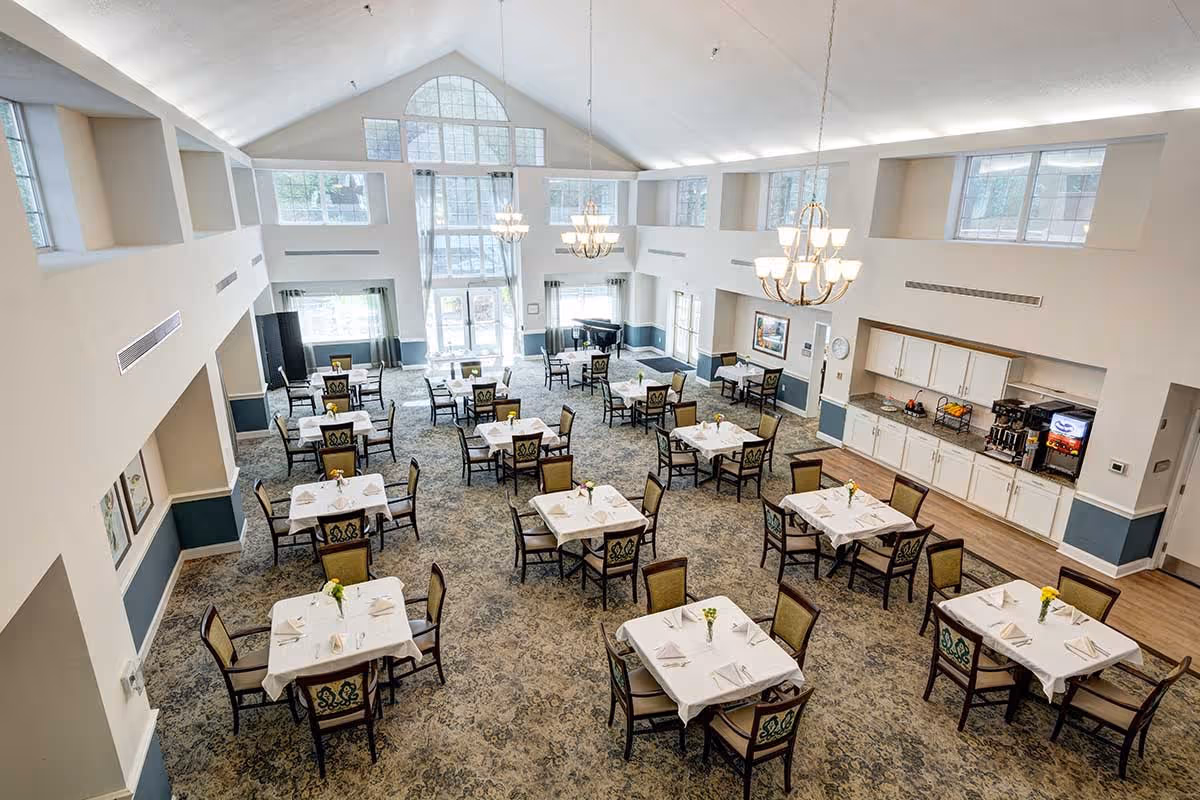 Spacious dining room with multiple square tables covered with white tablecloths, each set with napkins and small flower vases. The room has high vaulted ceilings, large windows letting in natural light, chandeliers, and a side area with cabinets and beverage machines.