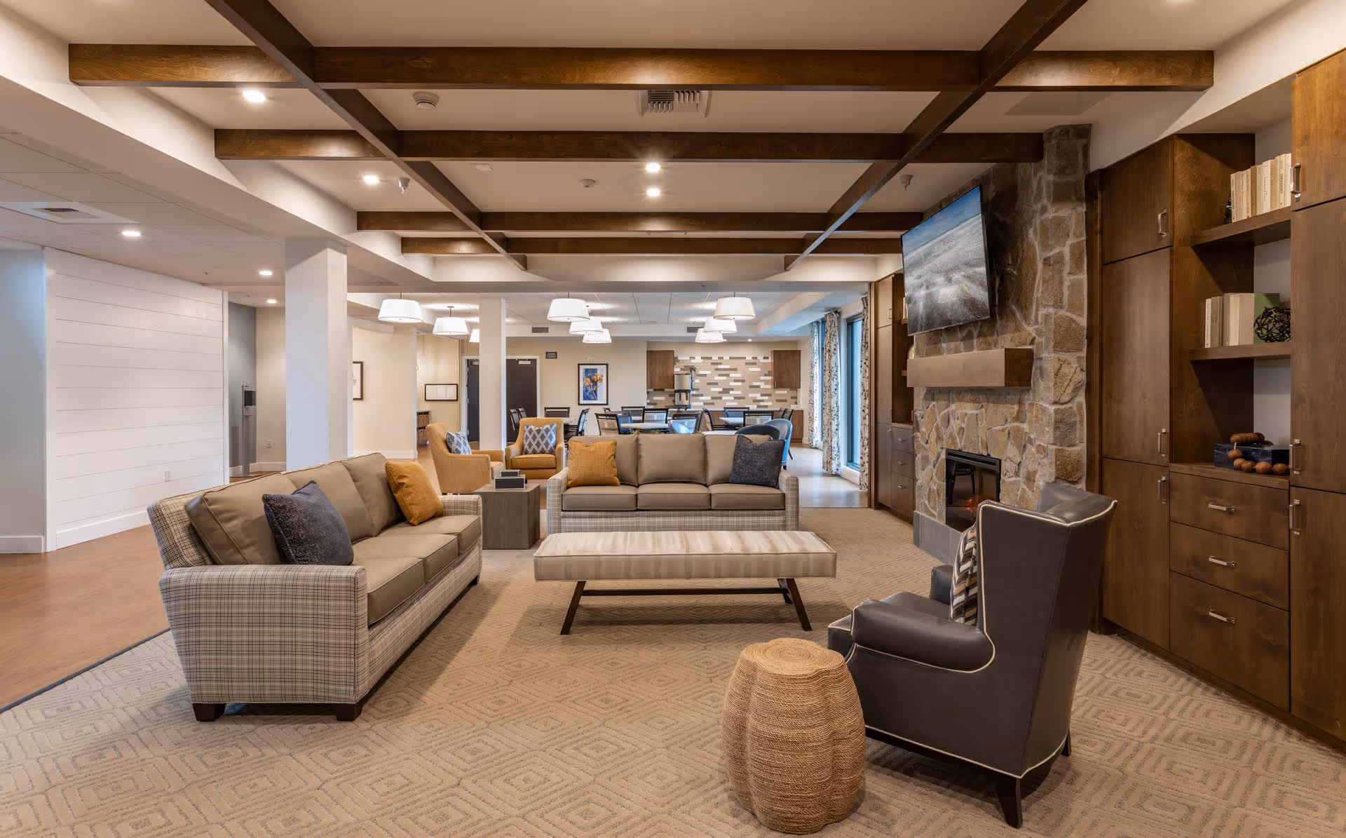 A spacious and well-lit living room area in a senior living facility featuring two beige sofas with accent pillows, a striped bench, a dark leather armchair with a patterned pillow, and a woven ottoman. The room has a stone fireplace with a mounted flat-screen TV above it, wooden built-in cabinets with books and decor, and exposed wooden ceiling beams. In the background, there is a dining area with tables and chairs, pendant lighting, and large windows with patterned curtains.