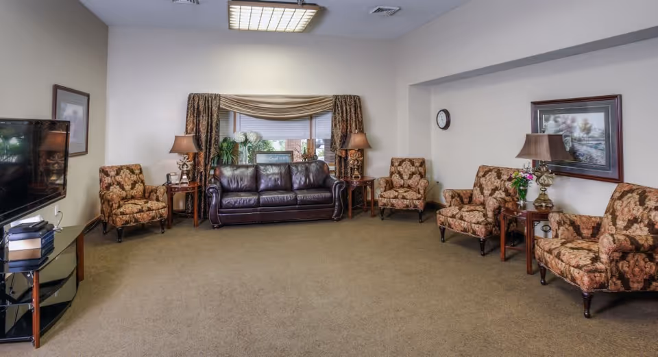 A spacious living room with a brown leather sofa centered under a window with drapes, flanked by two floral patterned armchairs and side tables with lamps. Additional matching armchairs and side tables with lamps and a framed picture are arranged along the right wall. A flat-screen TV sits on a glass stand on the left side of the room. The room has beige walls and carpeted floor.