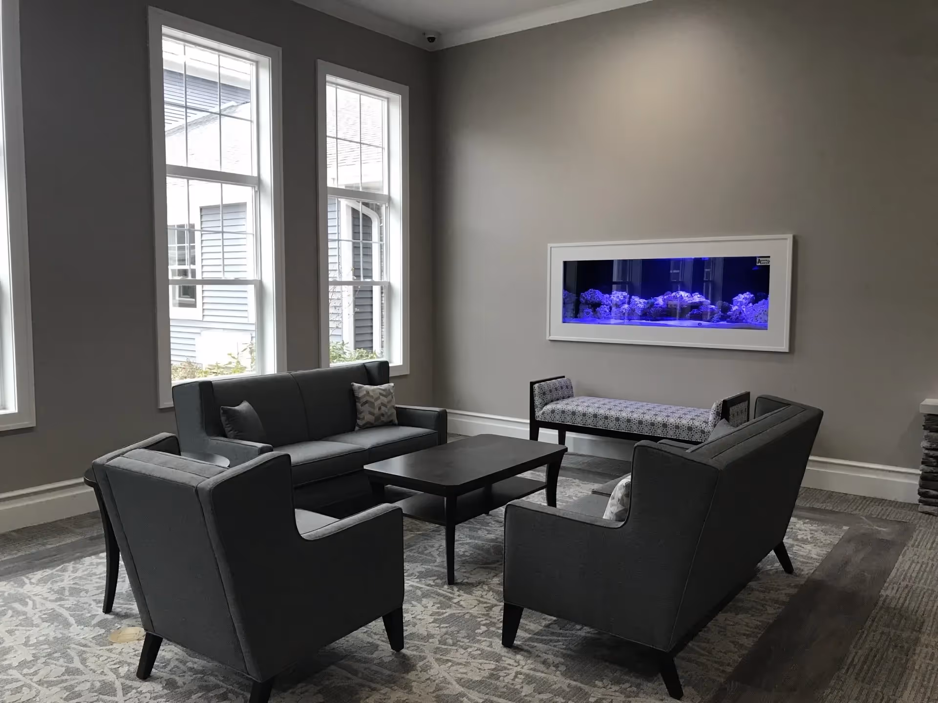 A modern senior living facility lounge area with gray upholstered sofas and armchairs arranged around a dark wooden coffee table. The room has large windows letting in natural light and a wall-mounted aquarium with blue lighting. A patterned bench is placed against the wall beneath the aquarium.