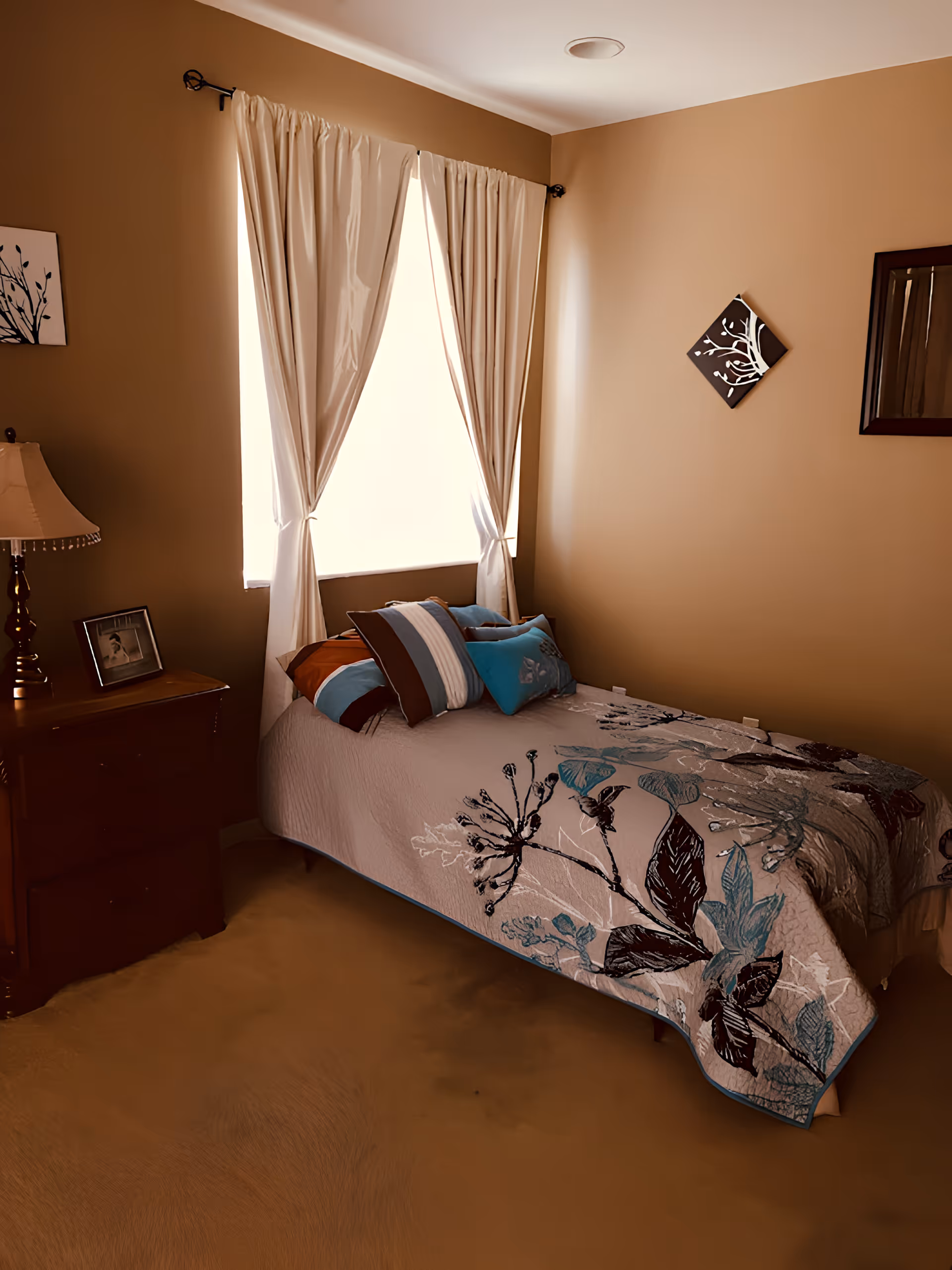 Small furnished bedroom with a single bed covered in a floral quilt, throw pillows, a nightstand with a lamp and a curtained window.