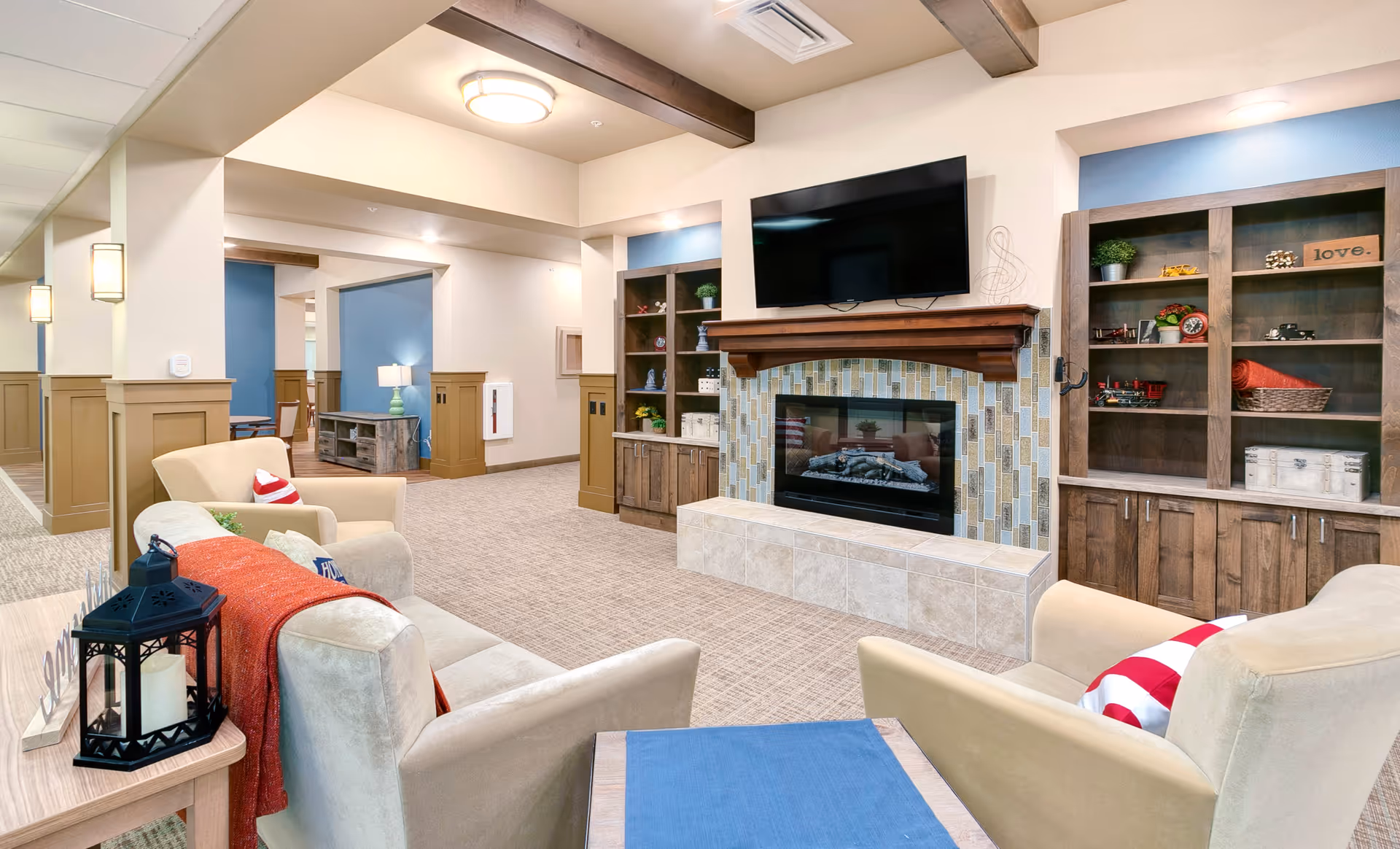 A cozy senior living common area with beige armchairs and a sofa arranged around a tiled fireplace with a mounted flat-screen TV above it. Wooden built-in shelves flank the fireplace, decorated with plants, books, and decorative items. The room features beige carpeting, wooden ceiling beams, and soft lighting, creating a warm and inviting atmosphere.