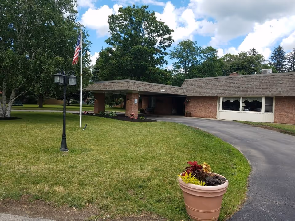 Exterior view of a single-story brick building with a covered entrance and a curved driveway. There is a well-maintained lawn with a large tree, a lamp post, an American flag on a flagpole, and a large flower pot with colorful plants in the foreground. The sky is partly cloudy.