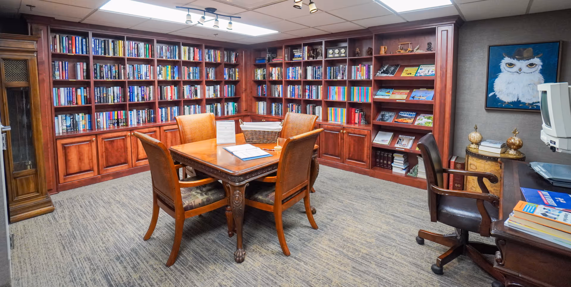 A cozy library room with wooden bookshelves filled with books lining the walls. In the center, there is a wooden table with four chairs around it. On the right side, there is a desk with a chair, some books, and a computer monitor. A framed picture of an owl wearing a hat hangs on the wall above a small decorative table with two golden ornaments.
