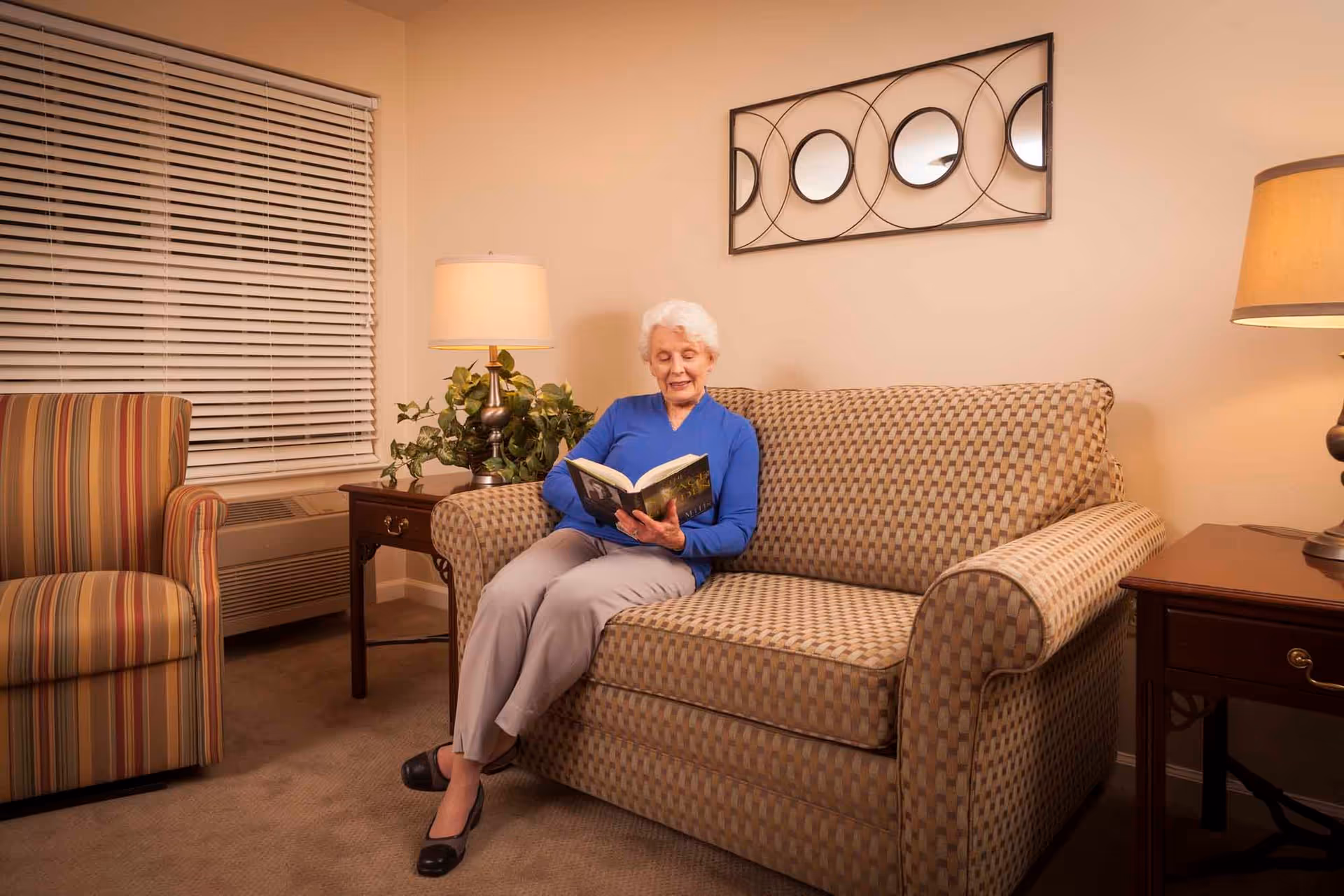 An elderly woman with white hair wearing a blue top and light-colored pants is sitting on a patterned sofa reading a book in a cozy living room. The room features a striped armchair, two wooden side tables with lamps, a potted plant, and a decorative wall mirror with circular designs. The window has closed blinds.
