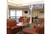 A cozy living room area with multiple cushioned chairs and sofas arranged around circular wooden tables. The room has beige walls, a carpeted floor, and a flat-screen TV mounted on the wall. There is a large window letting in natural light and some framed pictures and plants decorating the space.
